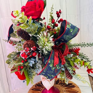 May include: A Christmas centerpiece featuring a Santa figurine with a red base, surrounded by greenery, red berries, pine cones, and a blue and green ribbon with a holly pattern.