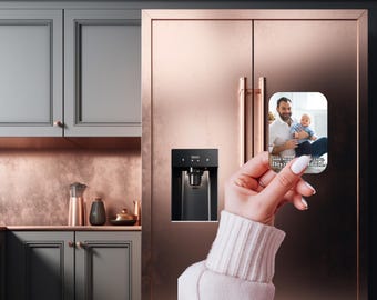 May include: A rectangular refrigerator magnet featuring a photo of a man holding a baby. The magnet is held in front of a rose gold refrigerator. Text on the magnet reads "Some heroes don't wear capes, they raise kids. Happy Father's Day Dad."