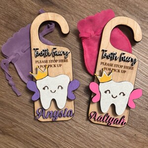 May include: Two wooden tooth fairy door hangers with a tooth fairy design, a crown, and wings. The hangers are personalized with the names "Anysia" and "Aaliyah". Each hanger has a small fabric bag attached.