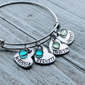 May include: A silver bangle bracelet with four heart-shaped charms. Each charm has a different name engraved on it: Maddox, Beckett, Emersyn, and Barrett. Each charm also has a small crystal stone.