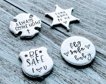 May include: Four silver metal tokens with different messages. The first token is a cross shape with the words "ALWAYS COME HOME" and a cross symbol. The second token is a star shape with the words "COME HOME TO ME" and a heart symbol. The third token is a shield shape with the words "BE SAFE I LOVE YOU" and a heart symbol. The fourth token is a circle shape with the words "FLY SAFE BABY" and a heart symbol.