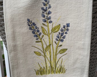 May include: A white kitchen towel with a floral design featuring purple lavender flowers and green leaves. The design is centered on the towel.