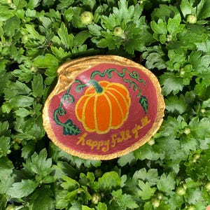 May include: A painted seashell with a gold rim, featuring a red background with a green vine and a pumpkin. The text "happy fall y'all" is written in gold lettering.