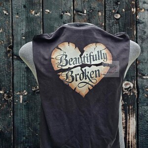 Peut inclure: D&eacute;bardeur noir avec un motif de c&oelig;ur vieilli. Le c&oelig;ur est divis&eacute; et porte les mots "Beautifully Broken" dans une police d&eacute;corative. Le haut est pr&eacute;sent&eacute; sur un mannequin.