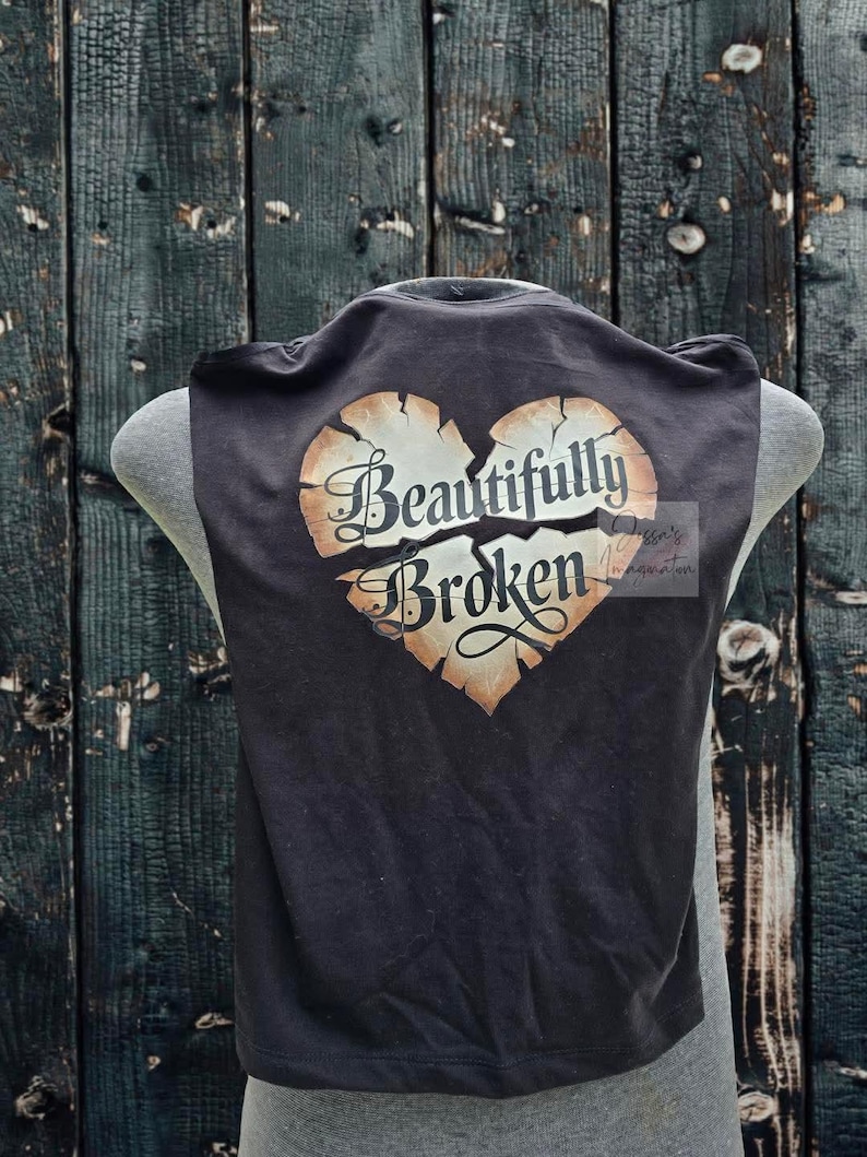 Peut inclure: D&eacute;bardeur noir avec un motif de c&oelig;ur vieilli. Le c&oelig;ur est divis&eacute; et porte les mots "Beautifully Broken" dans une police d&eacute;corative. Le haut est pr&eacute;sent&eacute; sur un mannequin.