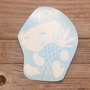 May include: White vinyl sticker of a cartoon character holding a pineapple. The character has a smiling face and a single antenna on its head.