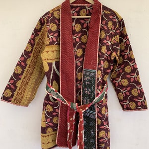 May include: A burgundy quilted robe with a floral pattern in yellow and pink. The robe has a matching belt and is displayed on a wooden hanger. The robe's design includes a mix of patterns and colours.