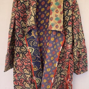 May include: A long, multi-coloured, patterned jacket with a shawl collar and a tie belt. The jacket is made of a patchwork of different fabrics, including floral prints and geometric patterns. The jacket is a warm, cosy, and stylish piece of clothing.