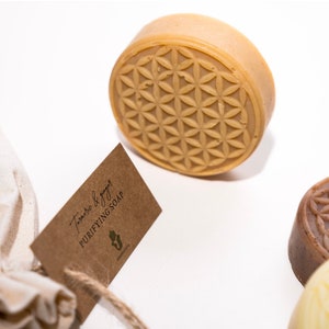 Puede incluir: Una pastilla redonda de jabón de cúrcuma y jengibre con un diseño de flor de la vida en la parte superior. El jabón está empaquetado en una pequeña bolsa de muselina blanca con una etiqueta de papel marrón que dice "Jabón purificante de cúrcuma y jengibre".