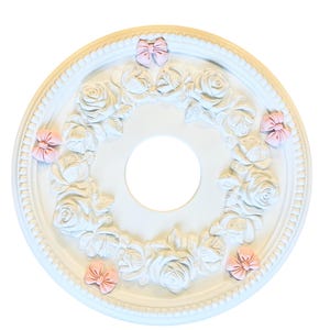 May include: A white, circular ceiling medallion with a central opening. The medallion features a raised floral design of roses and leaves, accented with small pink bows. The outer edge has a beaded trim.
