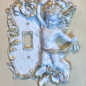 May include: White ceramic light switch cover with a gold cherub design. The cherub is holding a floral garland and has wings. The cover has a white background with a gold floral pattern.