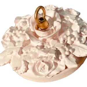 May include: A white ceramic ceiling fixture with a floral design and a gold metal hook. The fixture is shaped like a circle and has a raised design of roses and leaves.