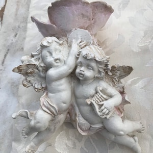May include: White ceramic wall decor featuring two cherubs, one playing a lyre and the other holding a round object. The cherubs are attached to a white flower-shaped base.