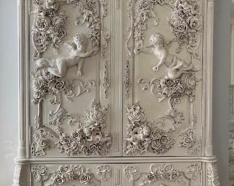 May include: A white, ornate, wooden armoire with intricate carvings of flowers and cherubs. The armoire has two doors and a base with a decorative design.
