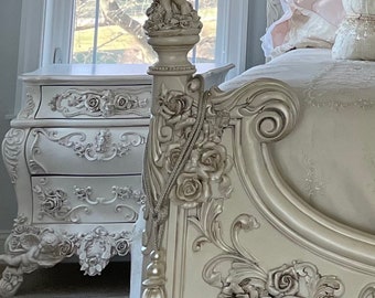 Puede incluir: Juego de muebles de dormitorio blancos ornamentados con una cama con tallas de rosas y querubines, una mesita de noche a juego y una cenefa decorativa. El marco de la cama tiene una cabecera y un piecero detallados. La habitación está decorada con un estilo clásico y elegante.