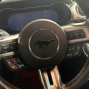 May include: A black leather-wrapped steering wheel with a silver trim and a black Mustang logo in the center. The steering wheel has controls for volume, navigation, phone calls, and music.