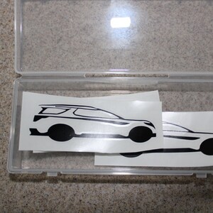 May include: Two black and white car silhouette decals in a clear plastic storage container. The decals are on white paper.