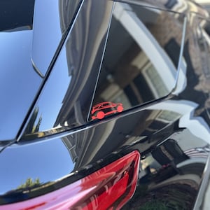 Peut inclure: Une voiture noire avec un autocollant rouge en forme de voiture sur la vitre arrière. La surface brillante de la voiture reflète l'environnement. Le feu arrière est rouge. L'autocollant est un contour stylisé d'un véhicule.