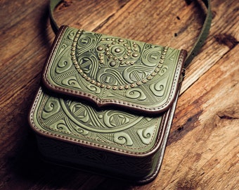 May include: A green leather crossbody bag with intricate embossed designs and gold-toned studs. The bag has a brown leather flap and a strap.