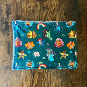 May include: A green fabric washcloth with a fringed border features a repeating pattern of Toy Story characters, including Woody, Buzz Lightyear, and Mr. and Mrs. Potato Head.