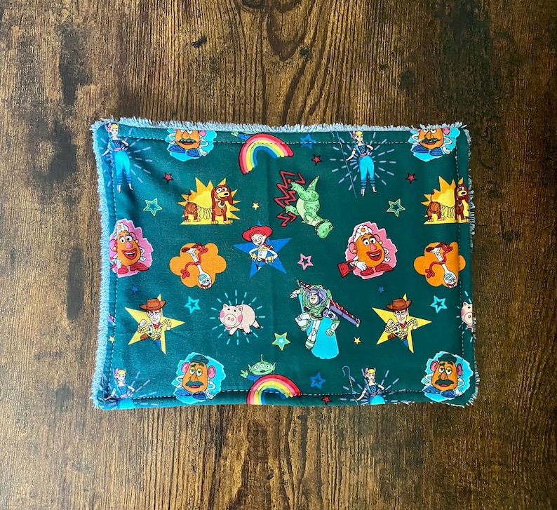 May include: A green fabric washcloth with a fringed border features a repeating pattern of Toy Story characters, including Woody, Buzz Lightyear, and Mr. and Mrs. Potato Head.