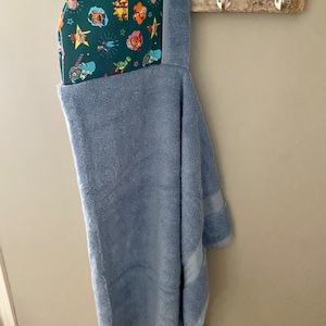 May include: A light blue hooded towel with a green and multi-colored lining featuring cartoon characters. The towel is hanging on a hook.