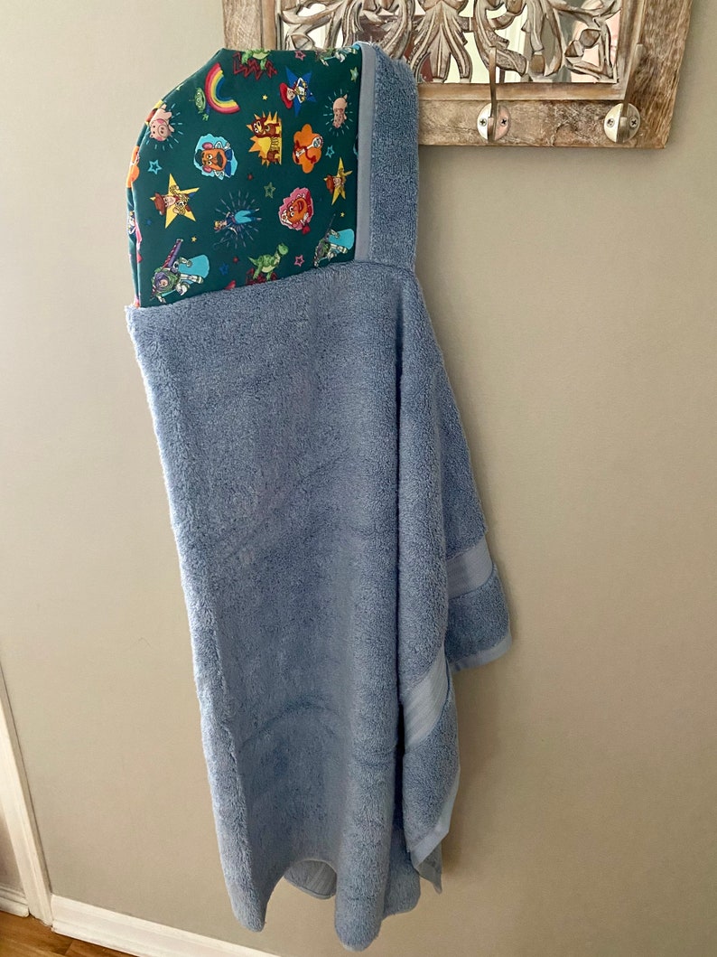 May include: A light blue hooded towel with a green and multi-colored lining featuring cartoon characters. The towel is hanging on a hook.