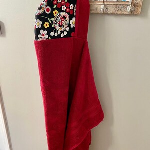 K&ouml;nnte beinhalten: Ein rotes Kapuzenhandtuch mit einem schwarzen und wei&szlig;en Blumendruck mit Minnie Mouse. Die Kapuze ist mit einem schwarzen und wei&szlig;en Blumenstoff gef&uuml;ttert.