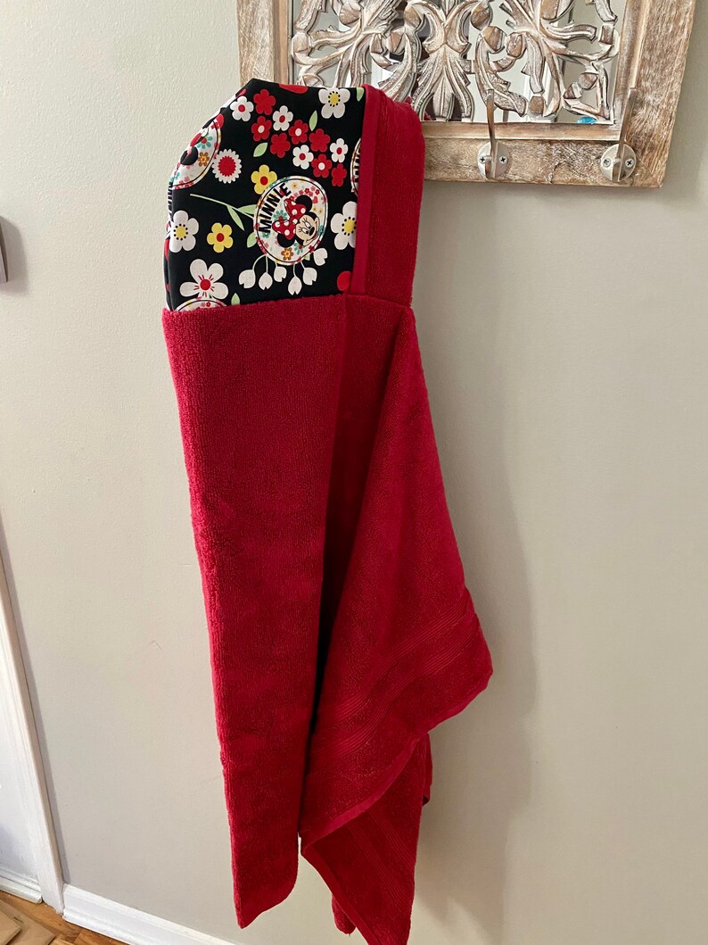 K&ouml;nnte beinhalten: Ein rotes Kapuzenhandtuch mit einem schwarzen und wei&szlig;en Blumendruck mit Minnie Mouse. Die Kapuze ist mit einem schwarzen und wei&szlig;en Blumenstoff gef&uuml;ttert.