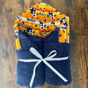 May include: A navy blue hooded towel with a white ribbon tied around it. The hood is made of a yellow and orange fabric with a white stripe pattern and the word "Astros" repeated throughout. The fabric also features a white "H" and stars.