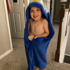May include: A young child wearing a blue hooded towel with a colorful pattern. The towel has a hood with a playful design. The child is smiling and looking at the camera.