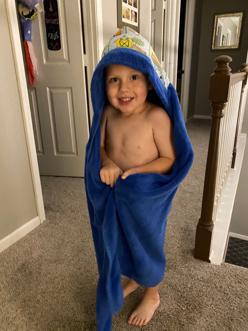 May include: A young child wearing a blue hooded towel with a colorful pattern. The towel has a hood with a playful design. The child is smiling and looking at the camera.