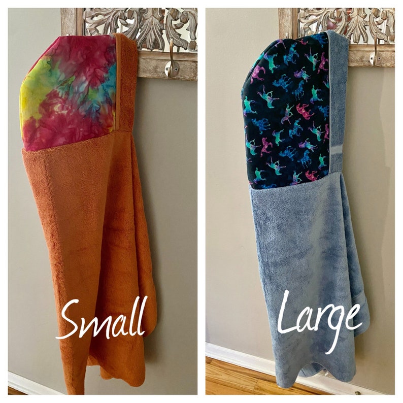 May include: Two hooded towels, one in a small size and one in a large size. The small towel is orange with a tie-dye pattern on the hood. The large towel is blue with a black and pink pattern of horses on the hood. Both towels are hanging on hooks.