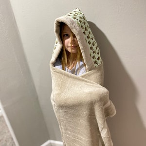 May include: A child wearing a hooded towel with a green and white pattern of cartoon characters. The towel is beige and has a white trim.