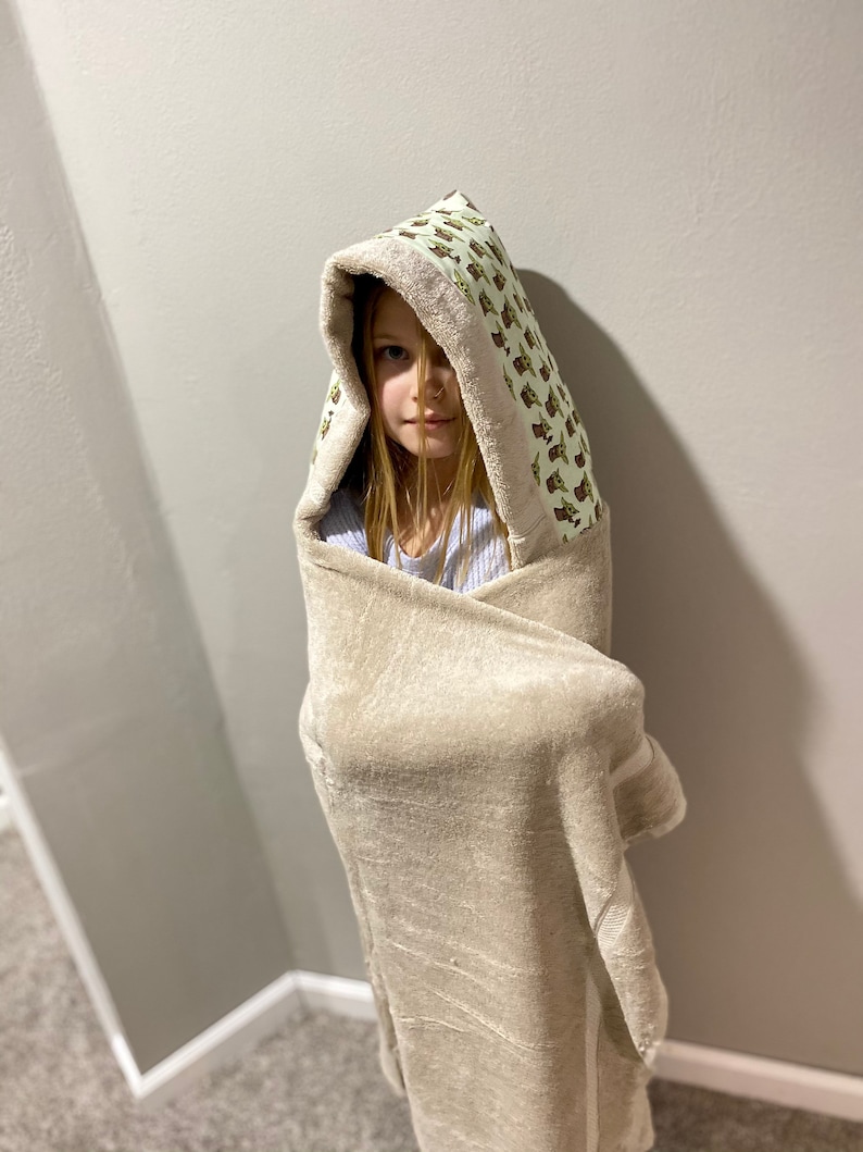 May include: A child wearing a hooded towel with a green and white pattern of cartoon characters. The towel is beige and has a white trim.