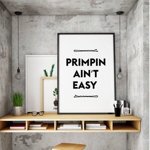 May include: A black and white framed print with the text "Primpin Ain't Easy" and two hair clips above and below the text. The print is hanging on a wall above a wooden shelf with books and glasses.