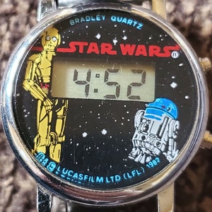 May include: A silver Star Wars digital watch with a black face. The face features C-3PO and R2-D2 characters, the words "Star Wars" and a digital display showing 4:52. The watch band is a silver metal link band.