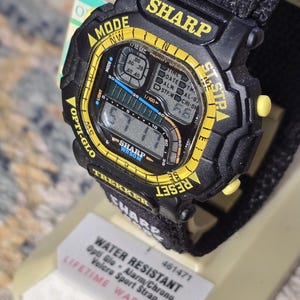 May include: Black and yellow Sharp digital watch with a black Velcro sport strap. The watch face displays the time and various functions. Features include Opti Glo, alarm, and chrono. The watch is water resistant.