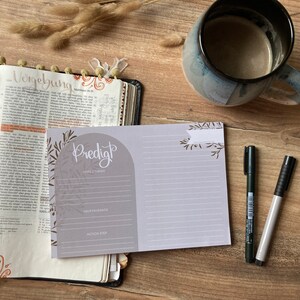 Puede incluir: Un diario gris y blanco con la palabra "Predict" escrita en cursiva marrón. El diario tiene un interior forrado y una sección para "Scripture & Theme", "Highlights" y "Action Steps".