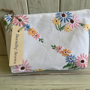 May include: A white fabric zip pouch featuring embroidered floral patterns in pink, blue, yellow, and green. A rectangular tan paper tag attached with twine displays the text Barnaby Brown in black ink on the front of the pouch.