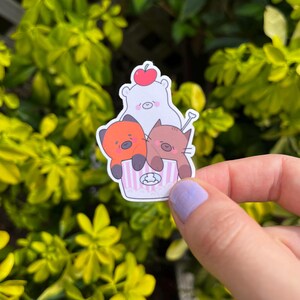 May include: A sticker featuring three cartoon animals: a white bear with a red cherry on its head, an orange bear, and a brown cat. They are in a pink and white striped cupcake wrapper. The sticker is held in front of a green leafy background.