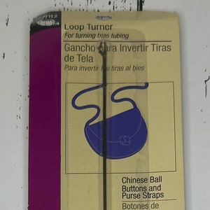 May include: A silver loop turner tool for turning bias tubing. The packaging shows illustrations of different uses for the tool, including turning straps for purses, frog closures, button loops, and shoulder straps. The packaging also includes the text "Loop Turner", "Gancho para Invertir Tiras de Tela", "Para invertir tiras al bies", "Size 9 1/2" (24.13cm)", "Chinese Ball Buttons and Purse Straps", "Botones de tela orientales y tiras para bolsos", "Frog Closures", "Cierres de rana", "Button Loops", "Presillas para botones", and "Shoulder Straps".