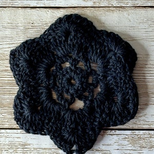 Puede incluir: Una flor de ganchillo negra con un contorno en forma de estrella. La flor está sobre un fondo de madera blanca. #4b