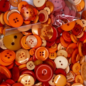 Op de afbeelding: Een grote verzameling oranje, bruine en crèmekleurige knopen in verschillende maten en vormen. De knopen zijn in een willekeurig patroon gerangschikt en zijn voornamelijk gemaakt van plastic.