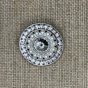 May include: A silver round button with a clear crystal centre surrounded by a ring of white pearls and a ring of clear rhinestones.