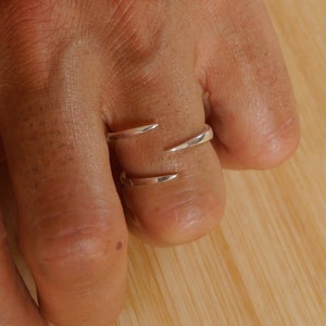 Könnte beinhalten: Ein silberner Ring mit zwei geschwungenen Bändern, die sich kreuzen. Der Ring wird an einem Finger getragen.