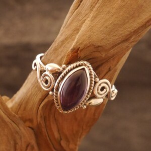 May include: A silver ring with an ornate design featuring a purple gemstone. The ring is set on a wooden surface.