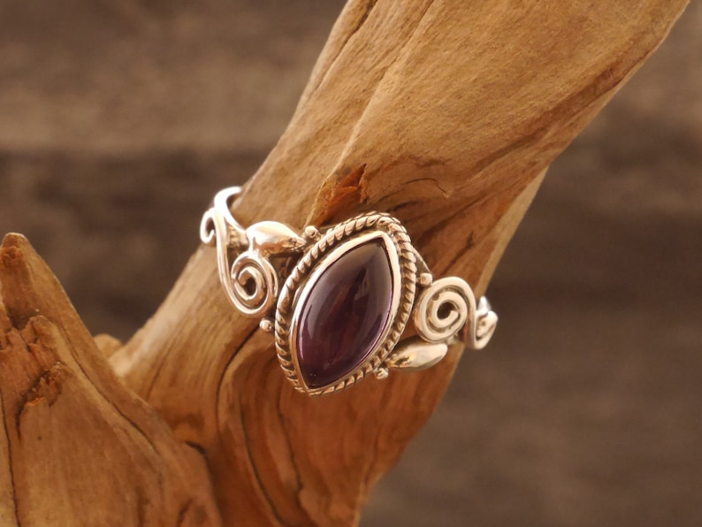 May include: A silver ring with an ornate design featuring a purple gemstone. The ring is set on a wooden surface.