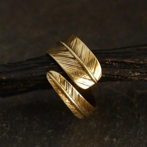 Könnte beinhalten: Ein goldfarbener Ring mit Federdesign. Der Ring hat zwei stilisierte Federn mit detaillierten Adern, die sich um den Finger wickeln. Der Ring ist vor einem dunklen, strukturierten Hintergrund platziert.