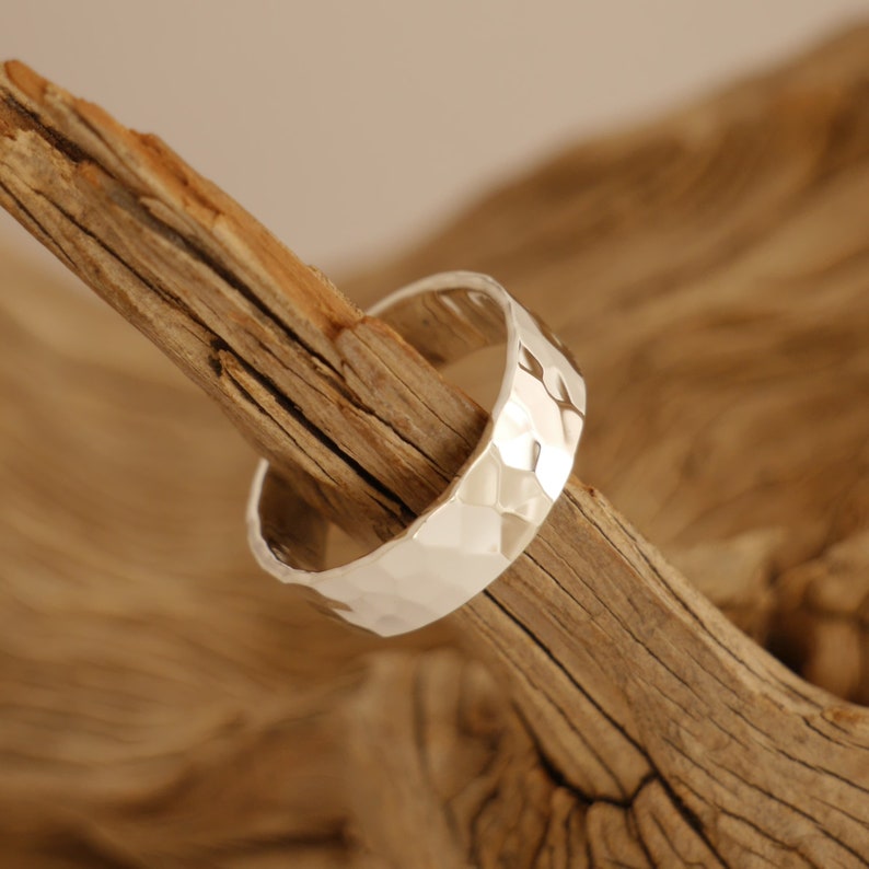 May include: A silver hammered band ring, displayed on a piece of driftwood.